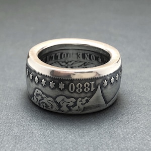 May include: A wide, silver ring crafted from a coin, featuring the year 1880 and star details. The ring has a polished finish and a detailed design, showcasing the coin's original engravings.