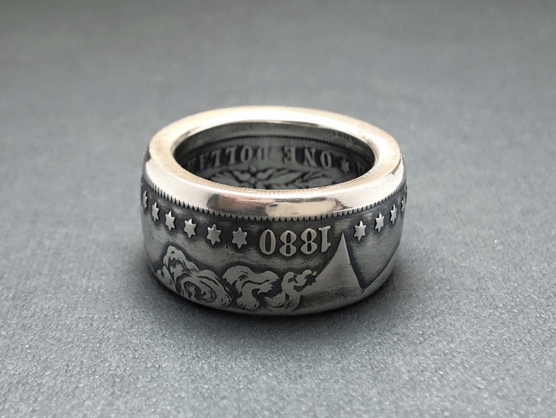 May include: A wide, silver ring crafted from a coin, featuring the year 1880 and star details. The ring has a polished finish and a detailed design, showcasing the coin's original engravings.