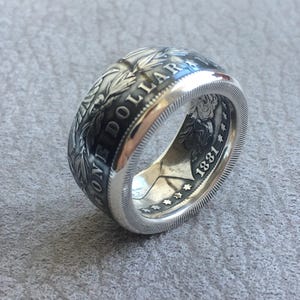 May include: A silver ring crafted from a coin, featuring intricate details and the inscription "ONE DOLLAR". The ring displays the year "1881" and a detailed design around the band, showcasing a vintage aesthetic.