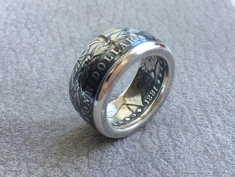 May include: A silver ring crafted from a coin, featuring intricate details and the inscription "ONE DOLLAR". The ring displays the year "1881" and a detailed design around the band, showcasing a vintage aesthetic.