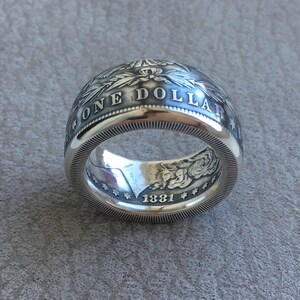 May include: A silver ring crafted from a coin, featuring the words "ONE DOLLAR" and the year "1881". The ring has intricate detailing and a polished finish, showcasing a vintage aesthetic.