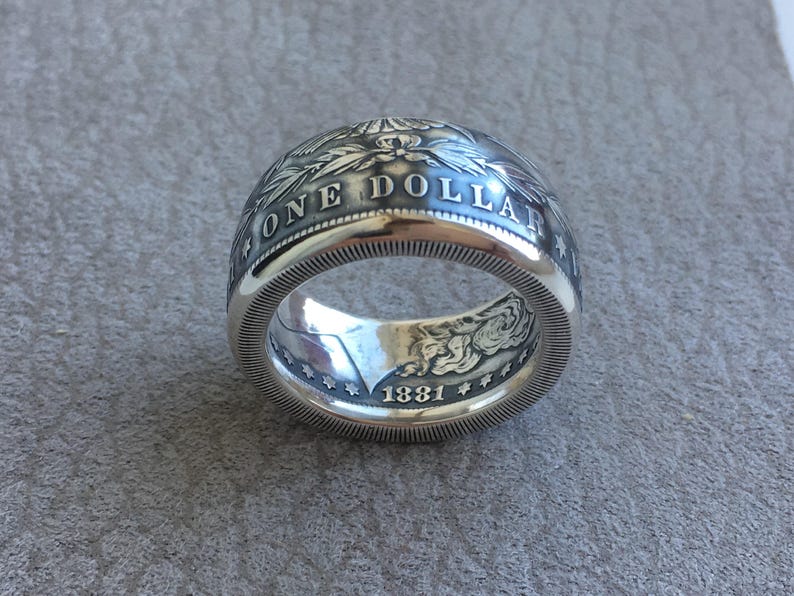 May include: A silver ring crafted from a coin, featuring the words "ONE DOLLAR" and the year "1881". The ring has intricate detailing and a polished finish, showcasing a vintage aesthetic.