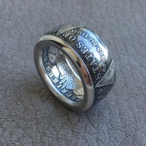 May include: A wide, silver ring with detailed engravings. The ring features text and imagery reminiscent of a US coin, including the words "STATES OF" and "GOD WE TRUST". The ring has a textured edge.