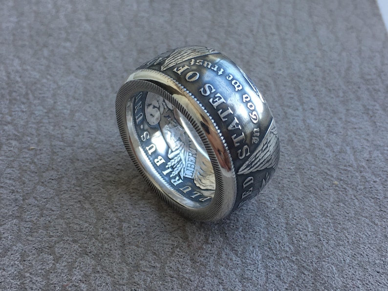 May include: A wide, silver ring with detailed engravings. The ring features text and imagery reminiscent of a US coin, including the words "STATES OF" and "GOD WE TRUST". The ring has a textured edge.