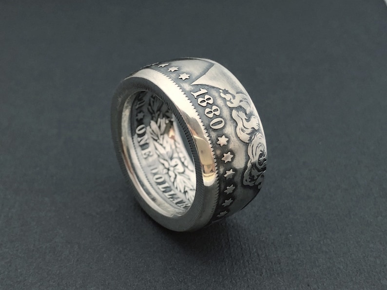 May include: A silver ring crafted from a coin, featuring the year 1880 and intricate details. The ring showcases a detailed design with stars and floral patterns, offering a unique and vintage aesthetic. The ring is a statement piece.