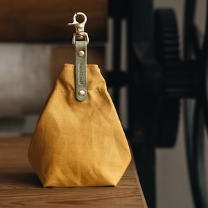 May include: A mustard yellow canvas pouch with a green leather strap and a gold metal clip. The pouch has the word "madewell" embossed on the strap.