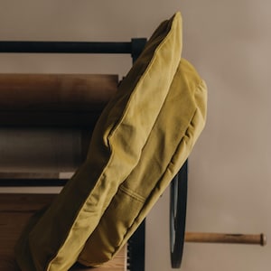 Pu&ograve; includere: Un cuscino giallo senape appoggiato su una superficie in legno. Il cuscino &egrave; realizzato in un tessuto morbido e testurizzato e ha una forma rettangolare semplice.