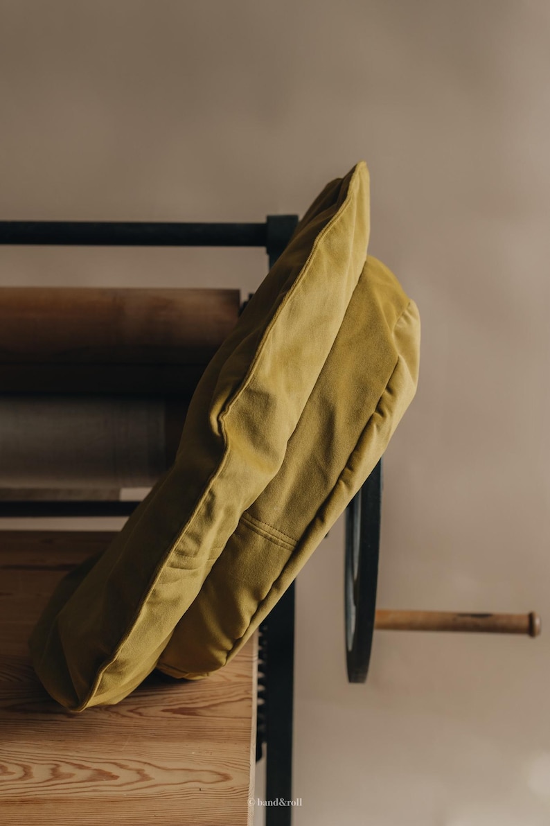 Pu&ograve; includere: Un cuscino giallo senape appoggiato su una superficie in legno. Il cuscino &egrave; realizzato in un tessuto morbido e testurizzato e ha una forma rettangolare semplice.
