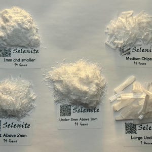 Może przedstawiać: Asortyment białych kryształów selenu w różnych rozmiarach, od 1 mm i mniejszych do dużych kawałków. Próbki są oznaczone informacjami o rozmiarze i wadze w gramach i uncjach. Kryształy mają różne tekstury.
