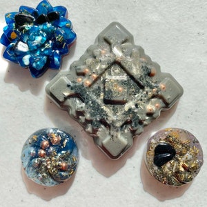 May include: Four resin ornaments with a blue, gray, and gold color scheme. The ornaments are shaped like a flower, a square, and two circles. The ornaments are filled with glitter, beads, and other decorative elements.