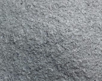 May include: Close-up of a pile of gray, crystalline flakes. The flakes have a slightly translucent appearance and vary in size, creating a textured surface. The overall color is a muted gray with subtle variations in tone.