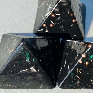 May include: Three black pyramid-shaped objects with gold and green glitter embedded in the resin. The pyramids are stacked on top of each other.