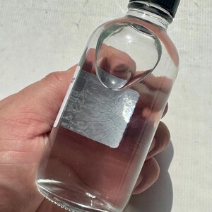 May include: A clear glass bottle with a black cap. The bottle has a unique, curved design on the side. The bottle is partially filled with a clear liquid. A silver label is affixed to the side of the bottle.