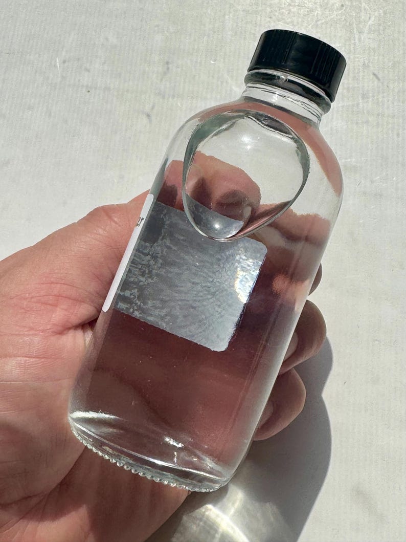 May include: A clear glass bottle with a black cap. The bottle has a unique, curved design on the side. The bottle is partially filled with a clear liquid. A silver label is affixed to the side of the bottle.