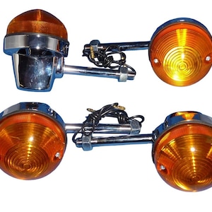 May include: Set of four motorcycle indicator lights. Each light has a chrome base and a clear amber lens. The lights are connected with metal rods and wiring. The lenses have a circular pattern.