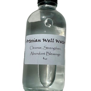 May include: A clear glass bottle with a black cap, filled with liquid. A white label on the bottle reads "Artesian Well Water" with the words "Cleanse, Strengthen, Abundant Blessings" and "4 oz".