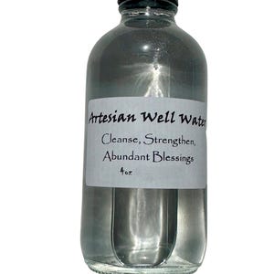May include: A clear glass bottle with a black cap, filled with a liquid. A white label on the bottle reads "Artesian Well Water, Cleanse, Strengthen, Abundant Blessings, 4 oz."