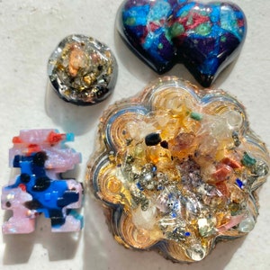 May include: Four resin-encased crystal charms, each with a unique design. The charms are a heart, a flower, a geometric shape, and a round shape. The charms are all different colors and have a variety of crystals embedded in them.