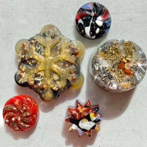 May include: Five resin ornaments with gold and silver glitter embedded in the resin. The ornaments are shaped like a snowflake, a circle, a star, and two swirls.