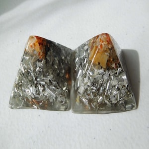 May include: Two clear crystal pyramids with silver flakes and orange and brown inclusions. The pyramids are sitting on a white surface.