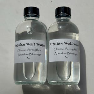 May include: Two clear glass bottles filled with water, each with a black cap. White labels on the bottles read "Artesian Well Water" with the words "Cleanse, Strengthen, Abundant Blessings" below. The bottles are 4 oz.
