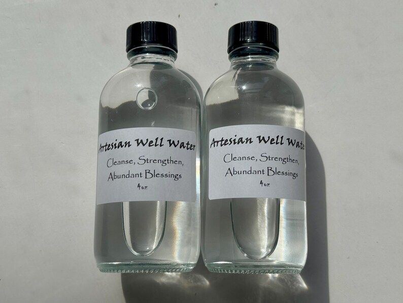 May include: Two clear glass bottles filled with water, each with a black cap. White labels on the bottles read "Artesian Well Water" with the words "Cleanse, Strengthen, Abundant Blessings" below. The bottles are 4 oz.