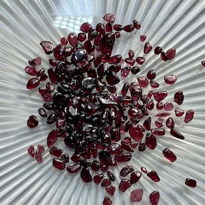 Op de afbeelding: Een close-up van een stapel kleine, donkerrode edelstenen op een wit, getextureerd oppervlak. De edelstenen zijn onregelmatig gevormd en hebben een glanzende afwerking.