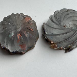 May include: Two decorative, swirled glass objects. The objects are gray with hints of copper and blue around the edges. The design is reminiscent of a spiral or a flower, with a textured surface.