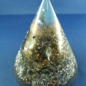 Pode incluir: Um cone de vidro transparente cheio de flocos metálicos prateados e cobre, pequenas contas de cobre e um cristal transparente. O cone está sobre um fundo azul.