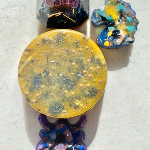 May include: Four resin ornaments in various shapes and colors. A yellow and black round ornament, a purple and blue unicorn head ornament, a purple and blue spider ornament, and a purple and blue geometric ornament.