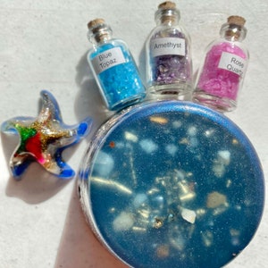 Può includere: Tre piccoli flaconi di vetro pieni di cristalli colorati. I flaconi sono etichettati "Blue Topaz", "Amethyst" e "Rose Quartz". I flaconi sono appoggiati su un sottobicchiere in resina blu con una stella marina.
