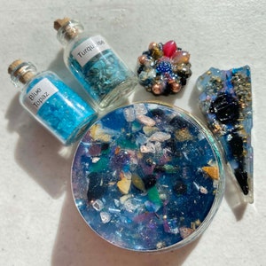 May include: A close-up of a round, blue resin coaster with embedded crystals and a variety of other materials. Two small glass bottles with blue and green crystals are labeled "Blue Topaz" and "Turquoise". A small, blue, gold, and red jeweled pin is also included.