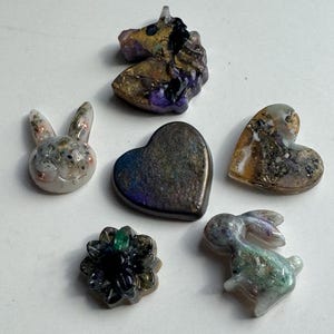 May include: A collection of small, resin-cast figurines in various shapes. Includes a unicorn, two rabbits, two hearts, and a flower. The figurines feature gold, purple, and green accents, with a glossy finish.