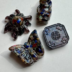 May include: Four decorative resin figurines: a spider, a gnome, a butterfly, and a square with an embossed design. The figurines are translucent with colorful inclusions of blue, red, and yellow. The spider has eight legs, and the butterfly has detailed wings.
