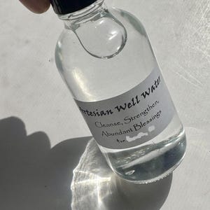 May include: A clear glass bottle with a black cap, filled with liquid. A label reads "Artesian Well Water, Cleanse, Strengthen, Abundant Blessings, 4 oz." The bottle is held at an angle, casting a shadow.