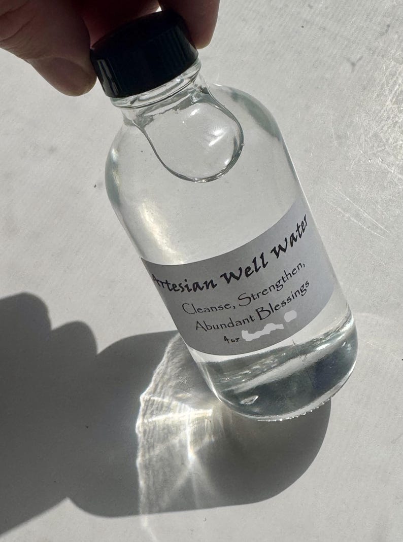 May include: A clear glass bottle with a black cap, filled with liquid. A label reads "Artesian Well Water, Cleanse, Strengthen, Abundant Blessings, 4 oz." The bottle is held at an angle, casting a shadow.