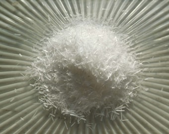 May include: A pile of white, thin, fibrous crystals on a textured, light gray surface.