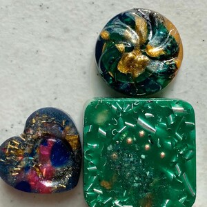 May include: Three resin coasters with different designs. One is a round green and gold swirl, one is a square green with silver and gold flecks, and one is a heart-shaped blue, pink, and gold design.
