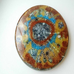 Puede incluir: Un objeto decorativo redondo, recubierto de resina, con un diseño circular de varios colores y texturas. El centro presenta un grupo de cristales claros y oscuros rodeados por un anillo de cuentas azules. El anillo exterior presenta una mezcla de cuentas y cristales dorados, marrones y blancos.