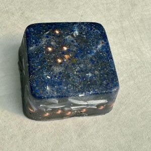 May include: A square, dark blue stone with copper-coloured flecks and accents. The stone has a speckled appearance and a dark, almost black, base. The top surface has a constellation-like pattern of copper-coloured dots.