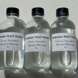 May include: Three clear glass bottles with black caps, each labeled "Artesian Well Water" with text below. The labels also read "Cleanse, Strengthen, Abundant Blessings" and "4 oz."
