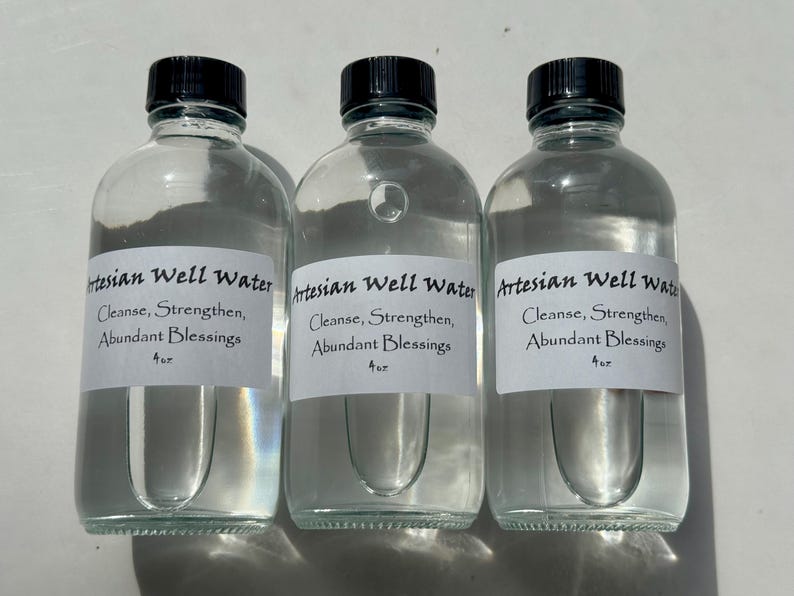 May include: Three clear glass bottles with black caps, each labeled "Artesian Well Water" with text below. The labels also read "Cleanse, Strengthen, Abundant Blessings" and "4 oz."