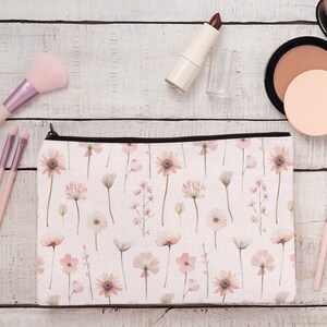 May include: A floral print makeup bag with a black zip, alongside makeup brushes, lipstick, and a compact. The bag displays a pattern of pink and white flowers on a light background. The makeup brushes have pink handles.
