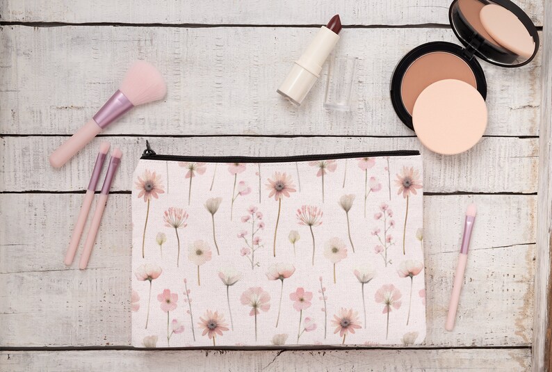 May include: A floral print makeup bag with a black zip, alongside makeup brushes, lipstick, and a compact. The bag displays a pattern of pink and white flowers on a light background. The makeup brushes have pink handles.