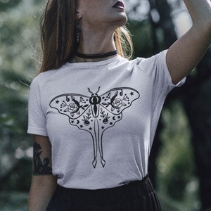 Może przedstawiać: Biały t-shirt z czarno-białym rysunkiem ćmy z kwiatowymi i niebieskimi detalami.