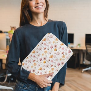 May include: A white laptop sleeve featuring a floral design in pink, peach, and yellow. The sleeve has a zip closure. The person is wearing a dark blue long-sleeved top and blue jeans.