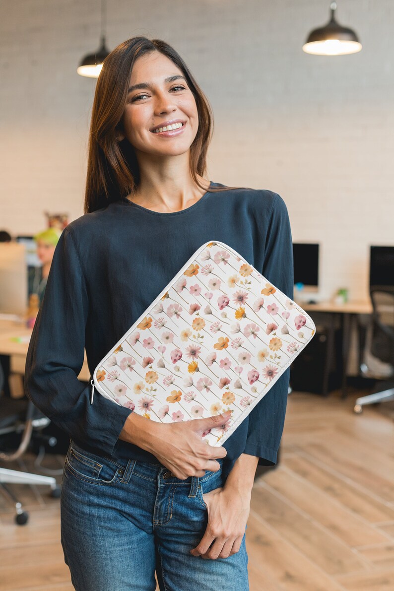 May include: A white laptop sleeve featuring a floral design in pink, peach, and yellow. The sleeve has a zip closure. The person is wearing a dark blue long-sleeved top and blue jeans.