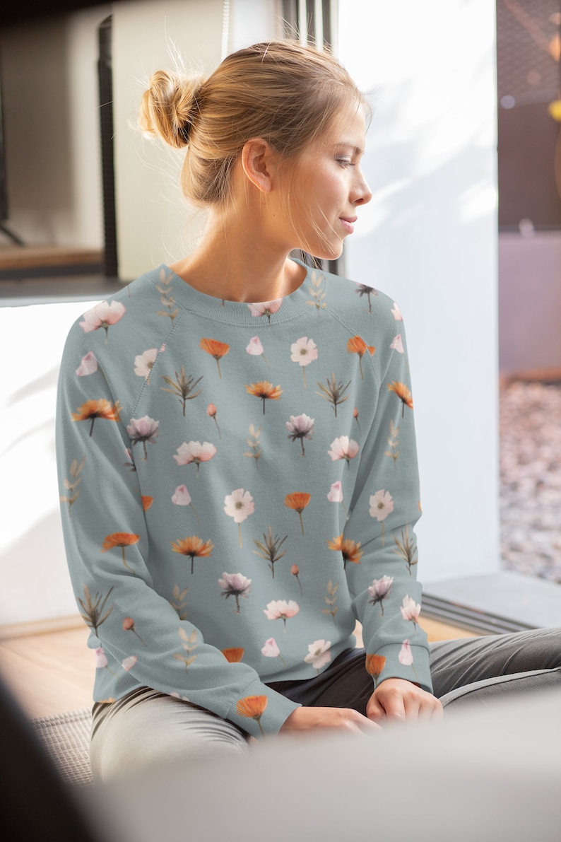 May include: A pale blue sweatshirt adorned with a repeating floral design of orange, white, and pink flowers, and green and brown leaves. The long-sleeved top is made of a soft material.