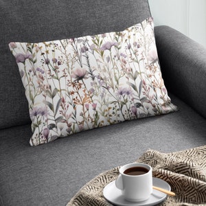 May include: Rectangular cushion with a floral design of purple, pink, and brown wildflowers on a white backdrop. The cushion is on a grey sofa with a blanket and a cup of coffee.