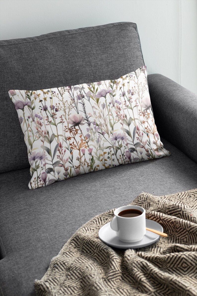May include: Rectangular cushion with a floral design of purple, pink, and brown wildflowers on a white backdrop. The cushion is on a grey sofa with a blanket and a cup of coffee.